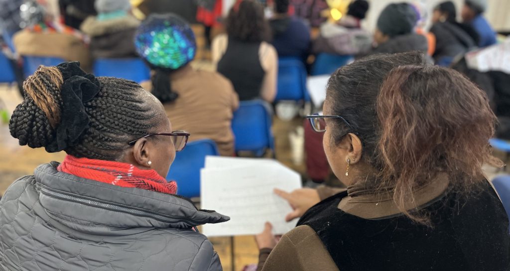 The backs of two women in a crowded hall, and the image behind them is of a blurred crowd